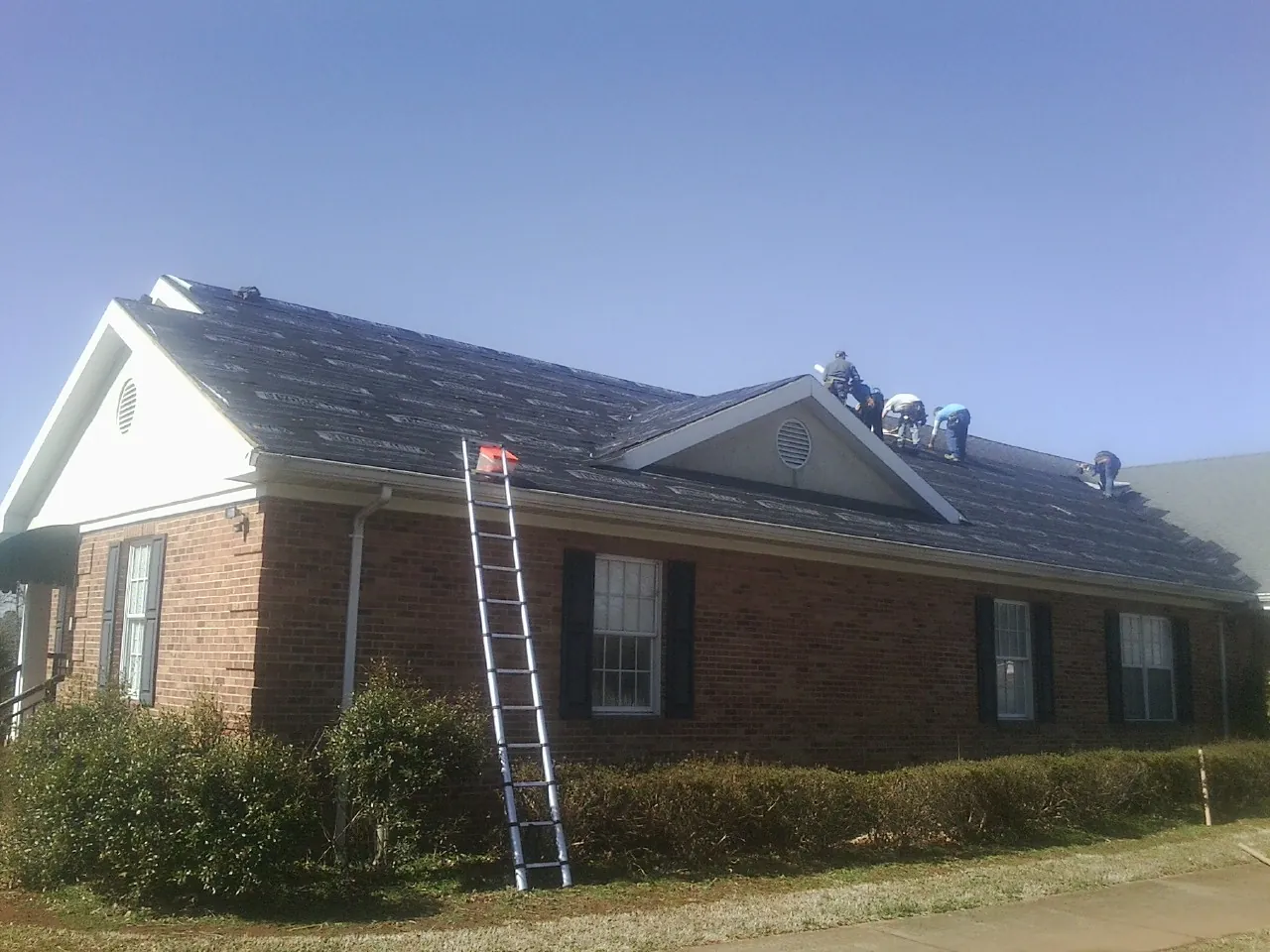 Crew tearing shingles off a brick ranch roof with a ladder set — Metal Roof Repair work by our crew in Lemon Springs