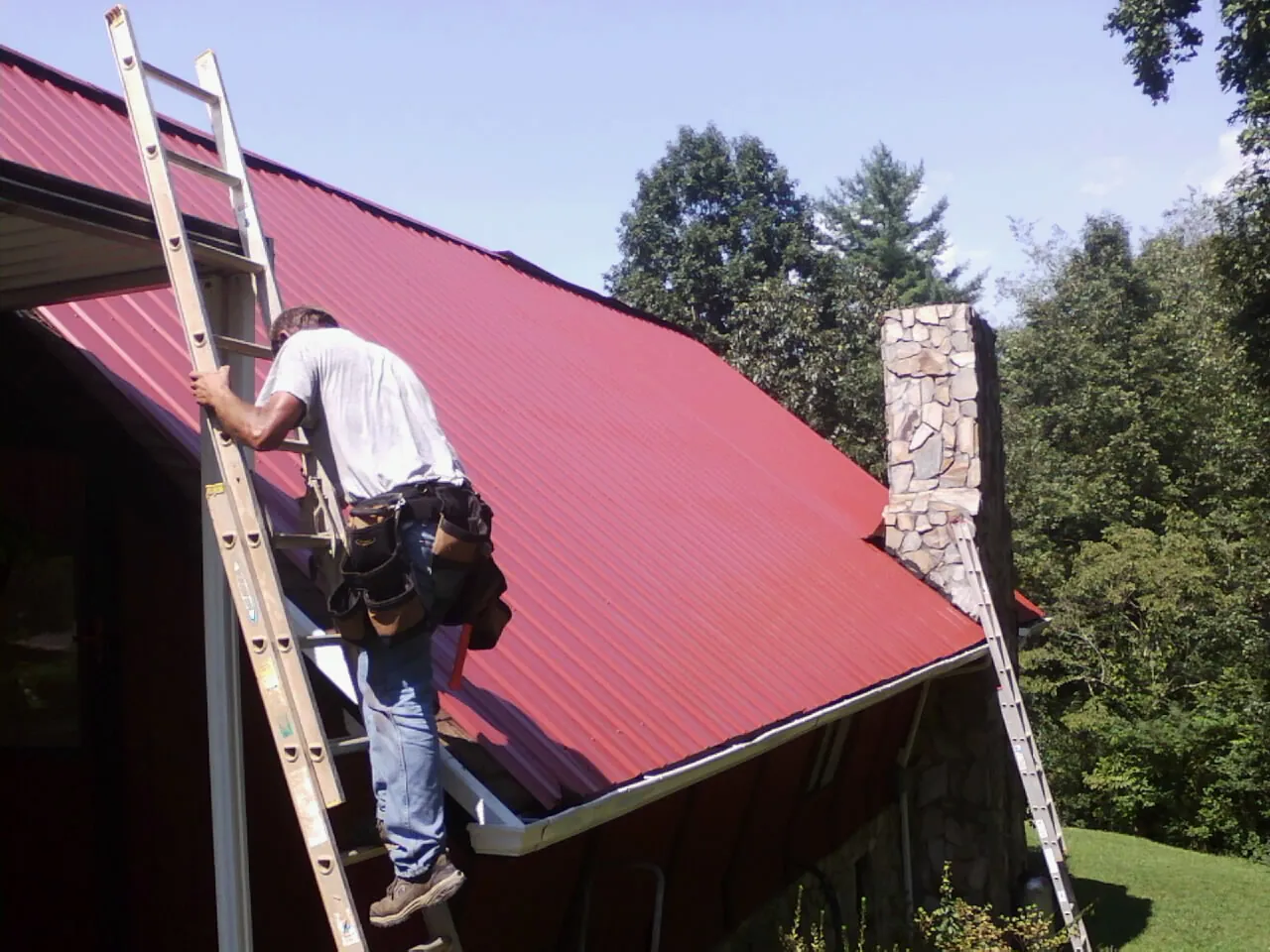 Roofer on a ladder beside a red metal roof and stone chimney — Soffit & Fascia Repair by Roofing Brothers in Lemon Springs